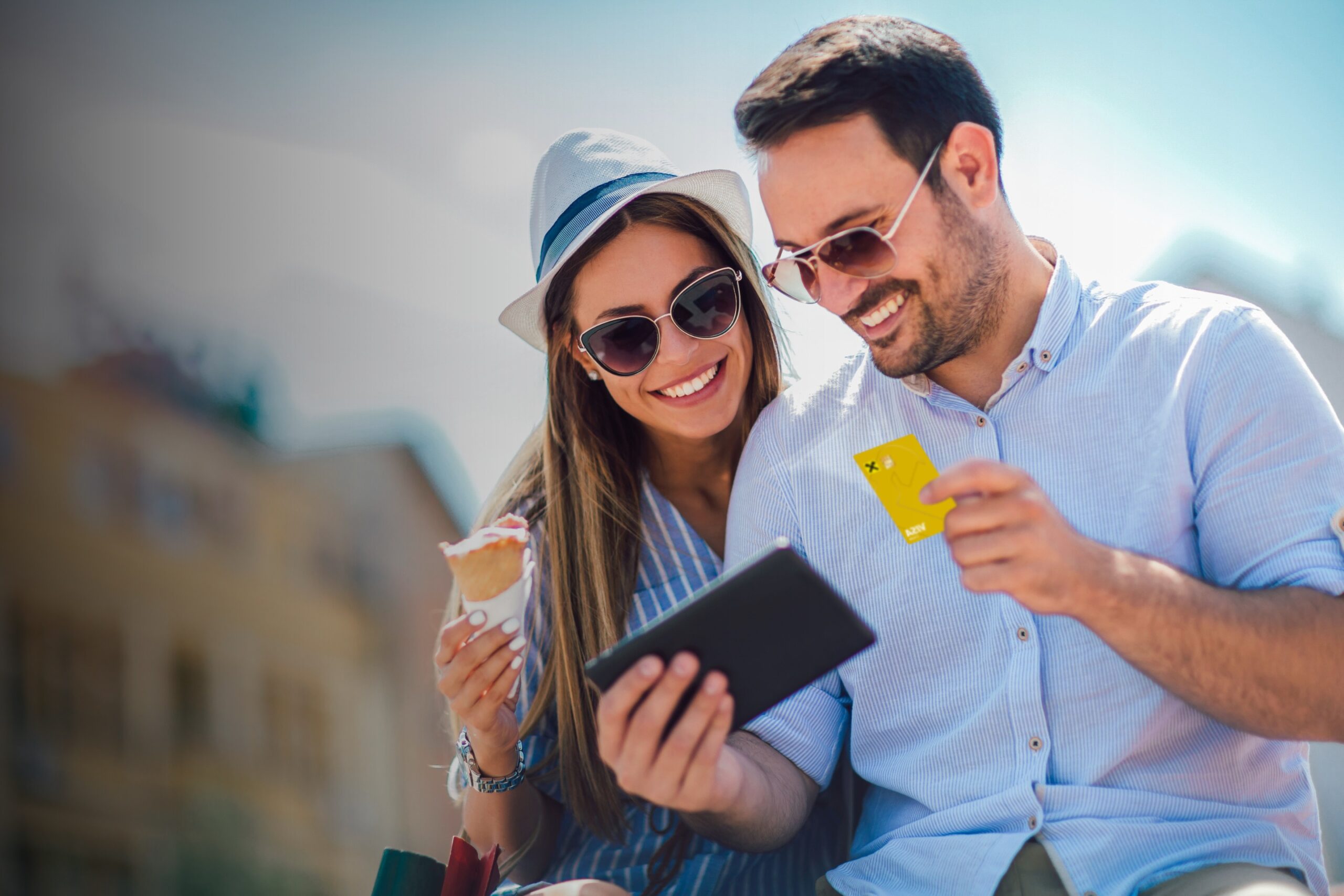 Paar im sommerlichen Outfit, die Frau ist ein Eis, der Mann hat die Raiffeisen Gold-Kreditkarte in der Hand, beide schauen auf ein kleines Tablet, das der Mann in der Hand hält